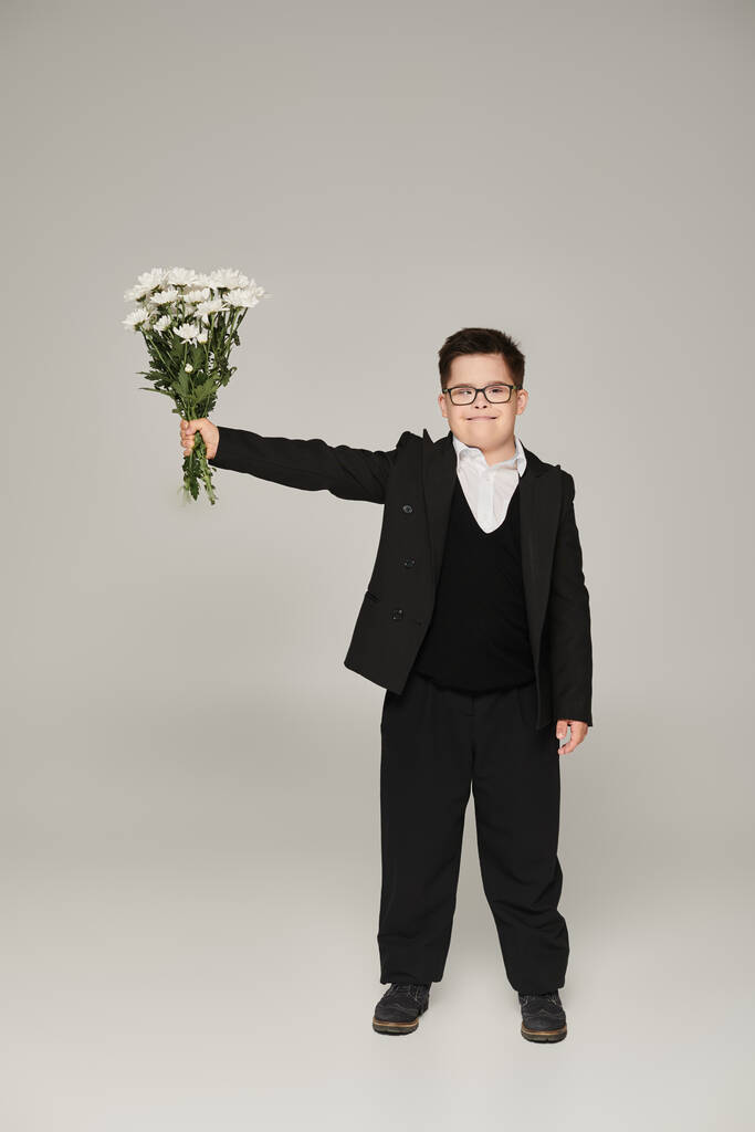 scolaro con sindrome di Down in occhiali e uniforme tenere bouquet in mano tesa su grigio - Foto, immagini