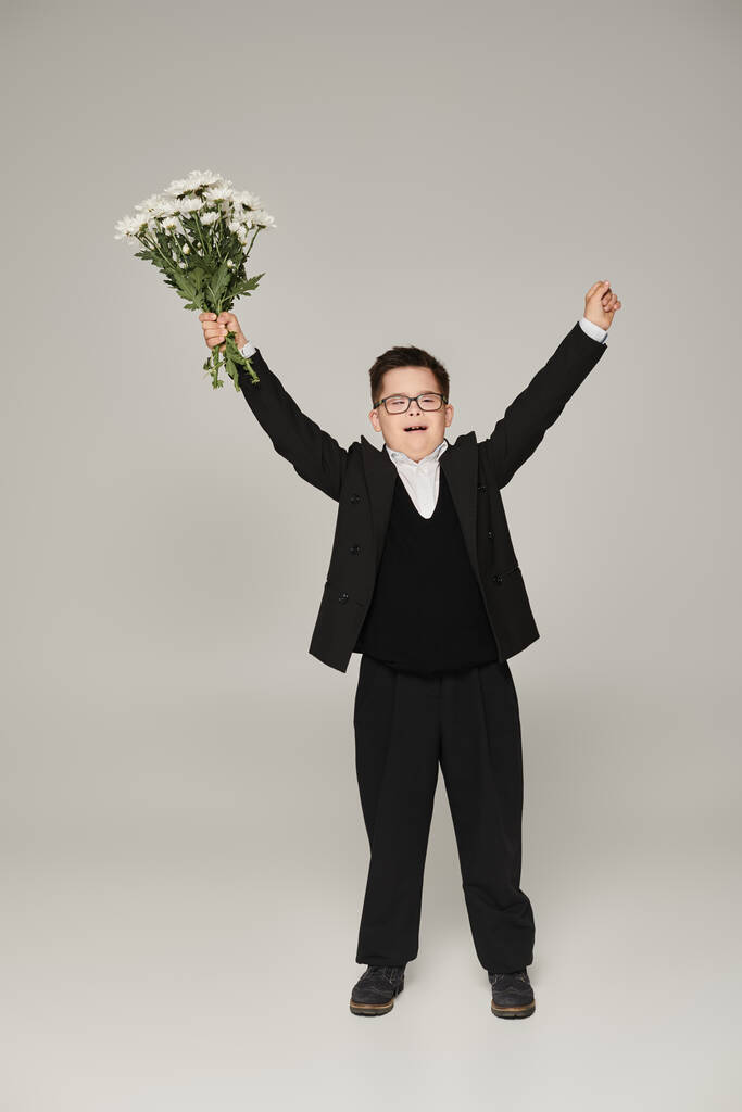 aufgeregter Schüler mit Down-Syndrom steht mit Blumen und erhobenen Händen auf grau, voller Länge - Foto, Bild