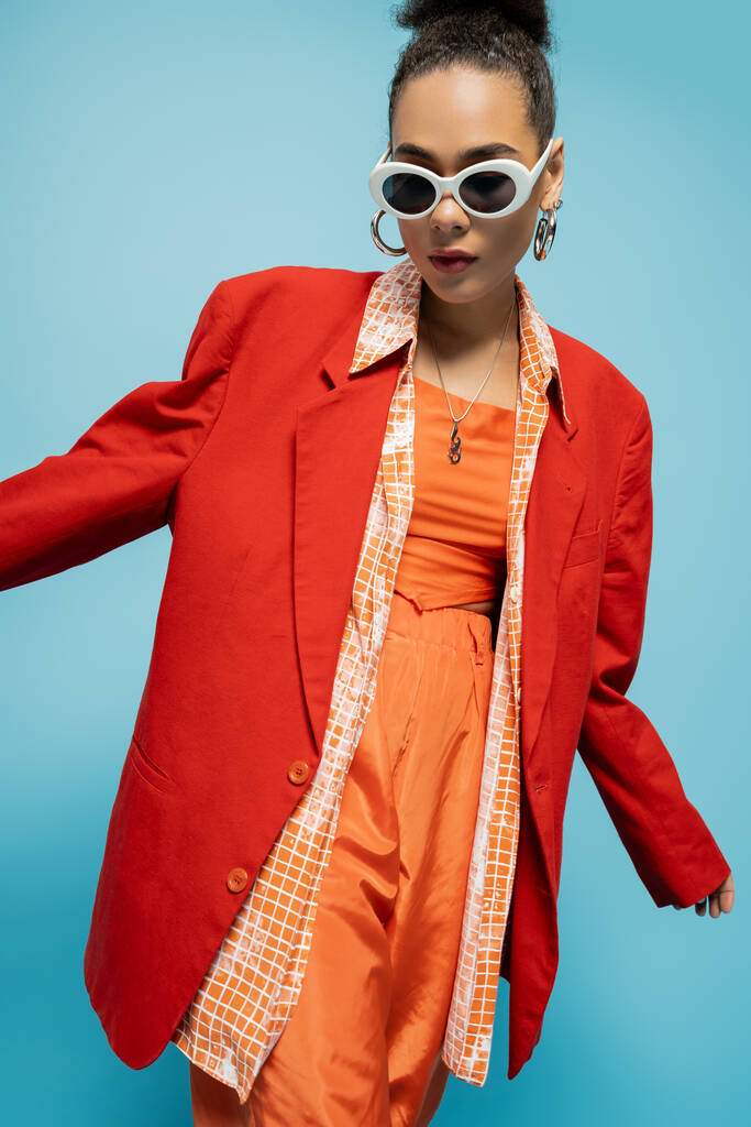 karizmatikus afro-amerikai nő divatos napszemüveg és élénk ruhát pózol a kék háttér - Fotó, kép