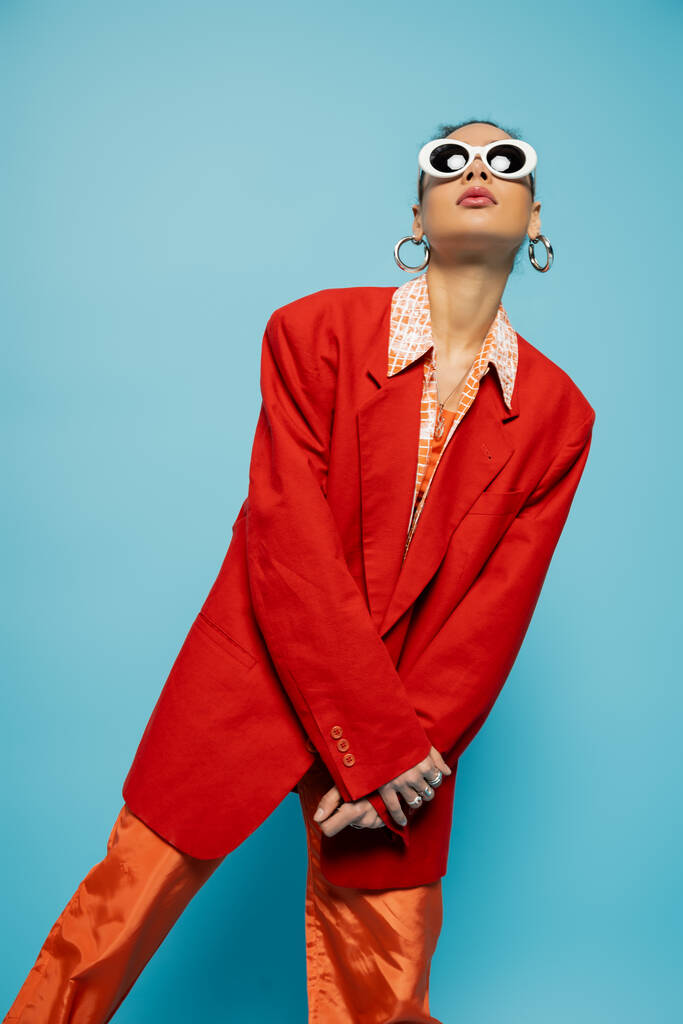 karizmatikus afro-amerikai nő divatos napszemüveg és merész ruhát pózol a kék háttér - Fotó, kép