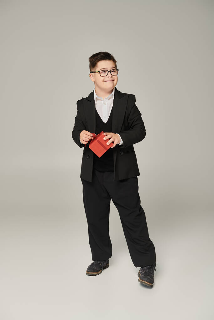 χαρούμενο αγόρι με σύνδρομο down στη σχολική στολή και γυαλιά ηλίου στέκεται με κόκκινο κουτί δώρου στο γκρι - Φωτογραφία, εικόνα