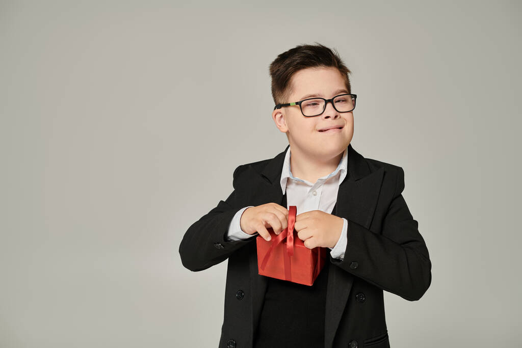 dziecko z zespołem Downa w okularach i mundurkach szkolnych z pudełkiem prezentów na szarym, specjalnym uczniu - Zdjęcie, obraz
