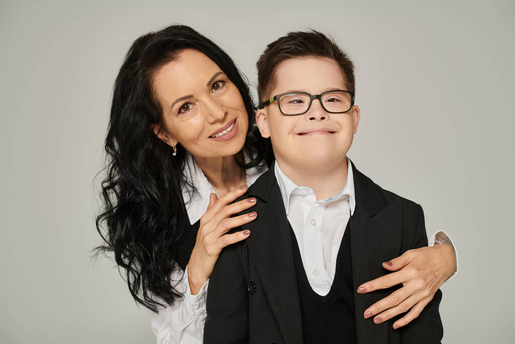 жінка середнього віку, що приймає сина з синдромом Дауна в шкільній формі, посміхаючись на камеру на сірому - Фото, зображення