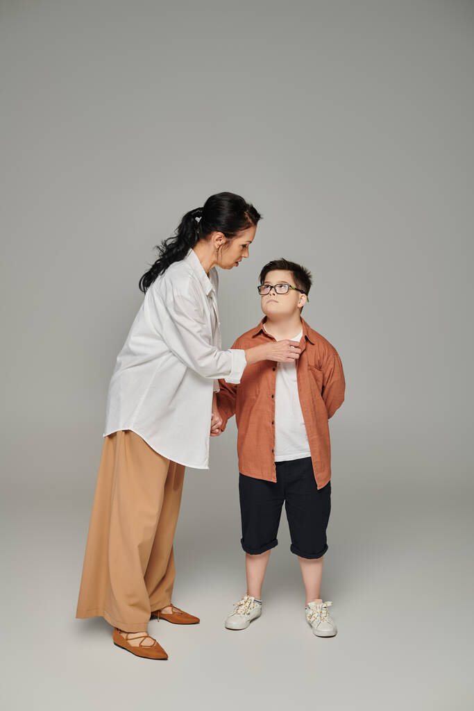 Женщина средних лет разговаривает с сыном с синдромом Дауна в очках на сером, поддерживающем родителе - Фото, изображение