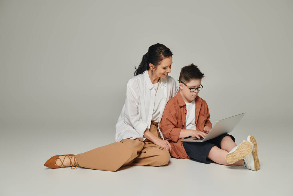 enfant atteint du syndrome du duvet assis près d'une mère souriante et utilisant un ordinateur portable sur gris, pleine longueur - Photo, image