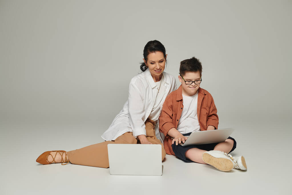 pozitivní žena středního věku a chlapec s Downovým syndromem sedí s notebooky na šedé, speciální rodiny - Fotografie, Obrázek