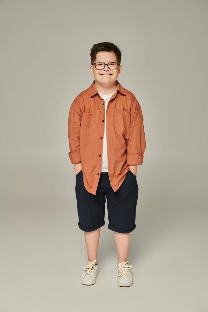 fröhlicher Junge mit Down-Syndrom in Shorts und Brille posiert mit den Händen in den Taschen auf grau - Foto, Bild