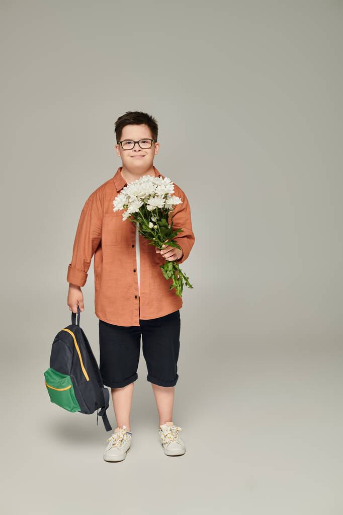 радостный мальчик с синдромом Дауна, в повседневной одежде и очках держа рюкзак и цветы на сером - Фото, изображение