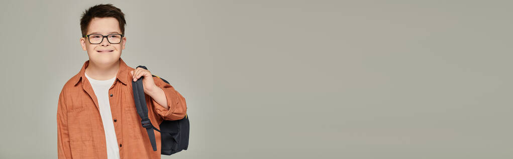 σχολιαρόπαιδο με σύνδρομο down σε κομψά ρούχα και γυαλιά κρατώντας σακίδιο πλάτης σε γκρι, banner - Φωτογραφία, εικόνα