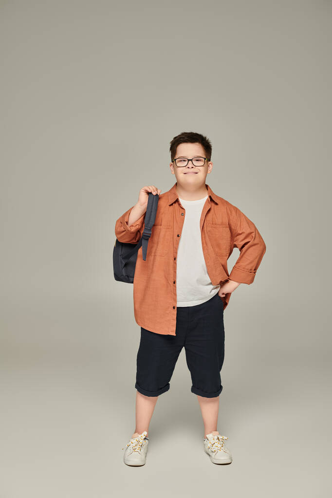 niño sonriente con síndrome de Down, en ropa de moda, posando con la mochila de la escuela en gris - Foto, Imagen