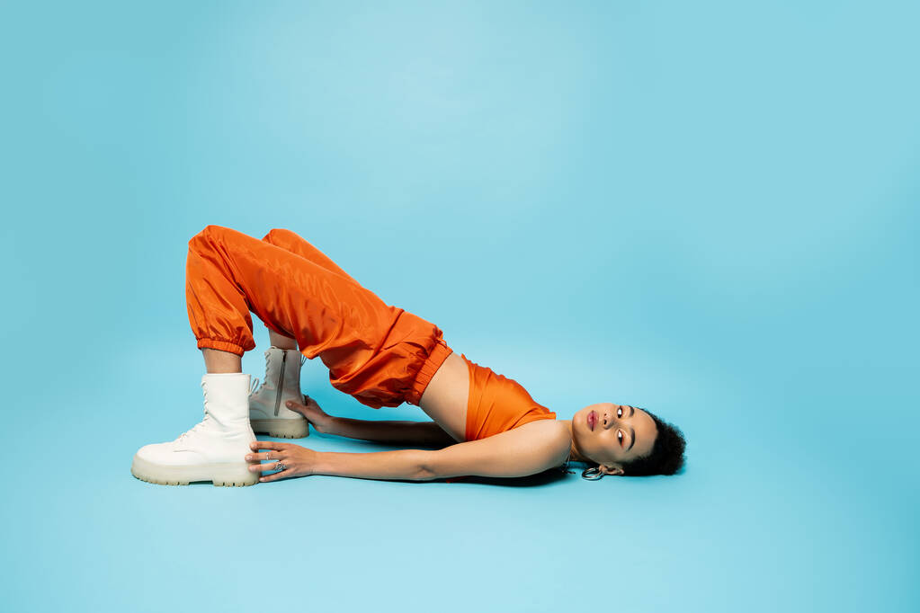 atrakcyjny model mody w żywe zużycie ulicy leżące na podłodze dotykając jej szpilki, niebieskie tło - Zdjęcie, obraz