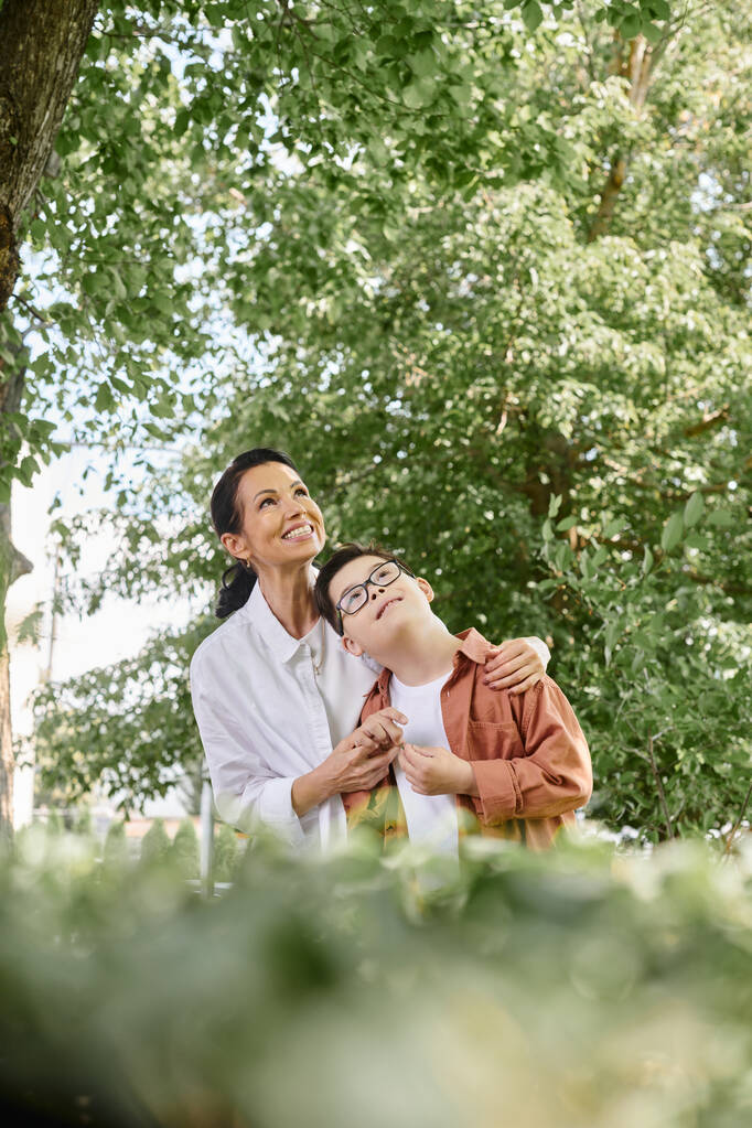 cheerful middle aged woman and son with down syndrome looking away on blurred foreground in park - Photo, Image