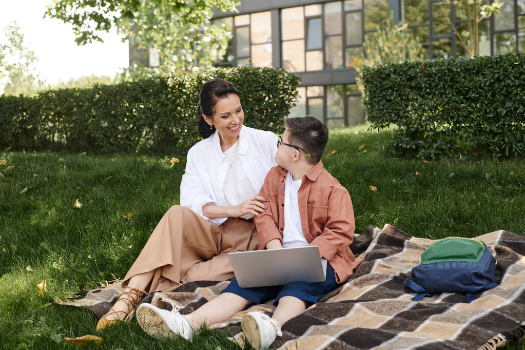 uśmiechnięta kobieta i chłopiec z zespołem Downa siedzi w pobliżu laptopa na kocu w parku, wyjątkowa rodzina - Zdjęcie, obraz