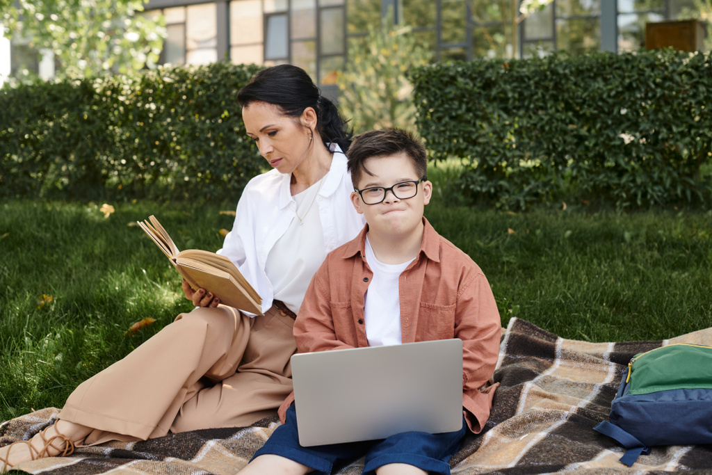 женщина средних лет читает книгу рядом с сыном с синдромом Дауна и ноутбук на одеяло в парке, досуг - Фото, изображение