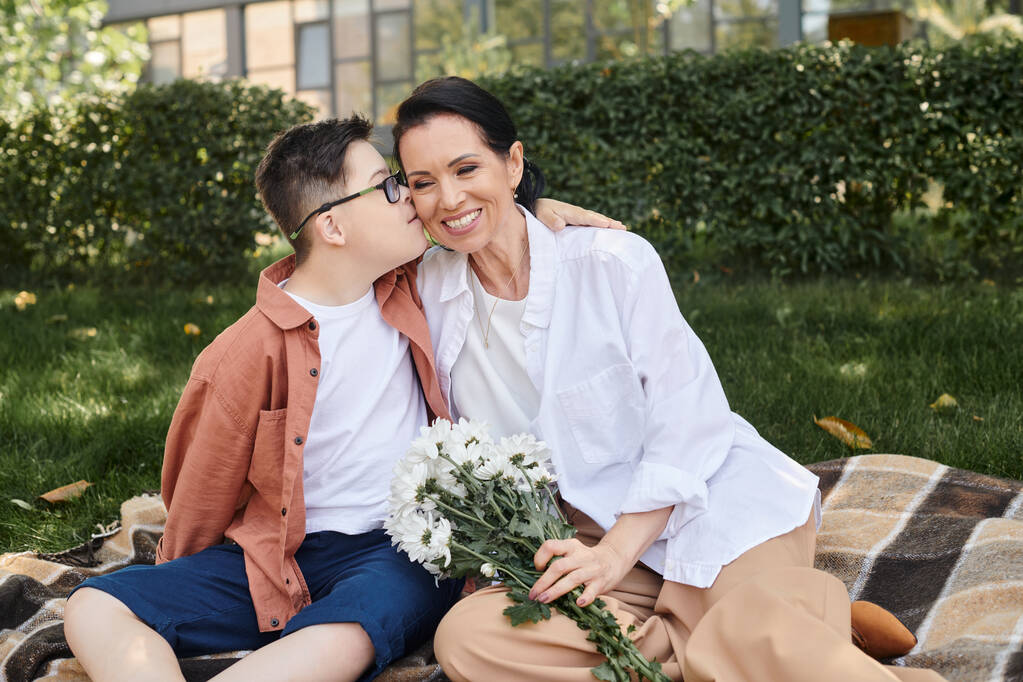 παιδί με σύνδρομο Down να φιλάει τη μητέρα του με λουλούδια σε κουβέρτα στο πάρκο, άνευ όρων αγάπη - Φωτογραφία, εικόνα
