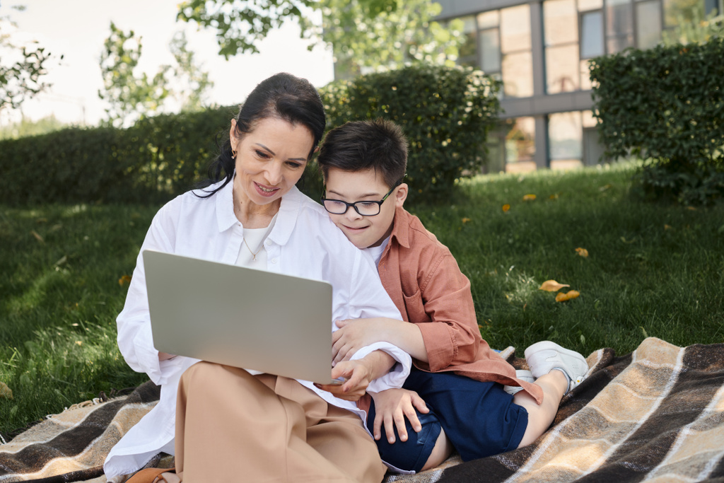 счастливая женщина средних лет сидит с ноутбуком рядом с сыном с синдромом Дауна на одеяле в парке - Фото, изображение