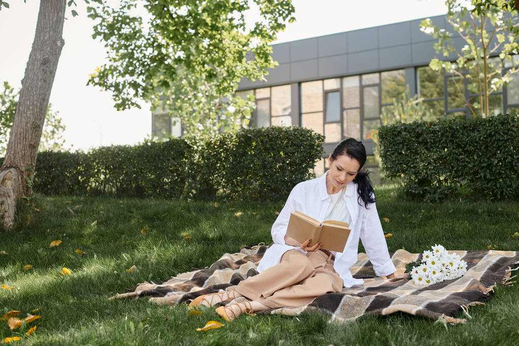 mujer de mediana edad en traje casual lectura novela mientras está sentado en manta en el parque, tiempo de calidad - Foto, imagen