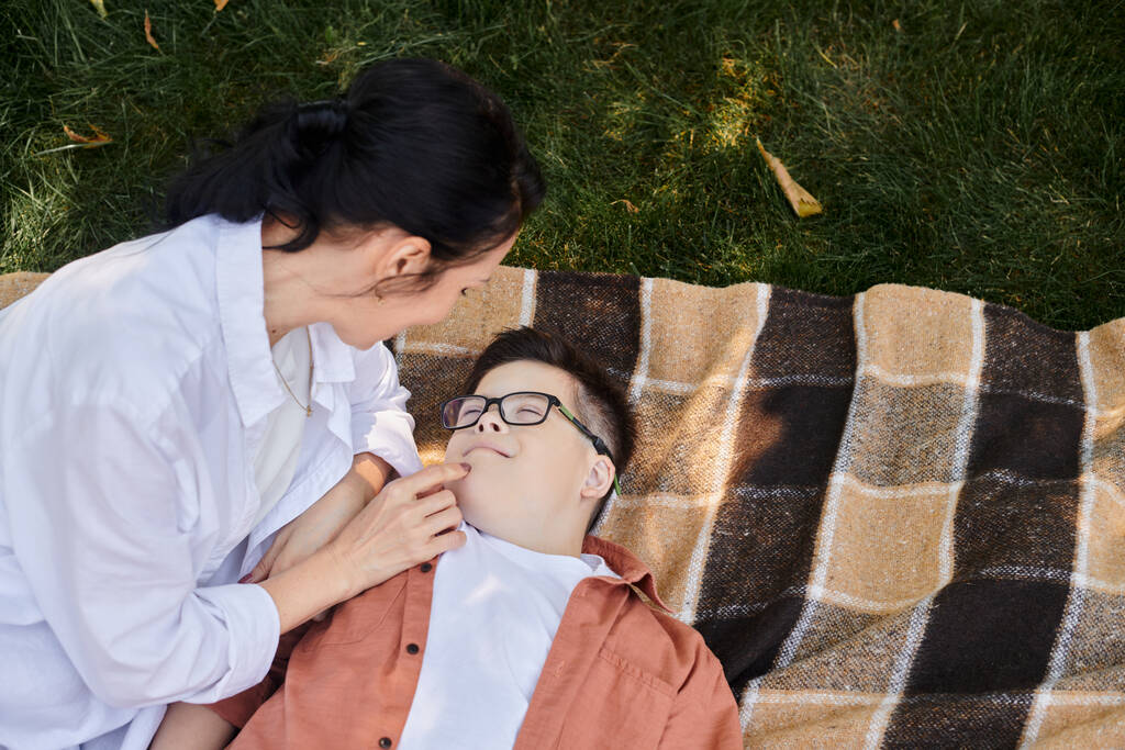 κορυφαία άποψη της γυναίκας αγγίζοντας το πρόσωπο του γιου με σύνδρομο κάτω στην κουβέρτα στο πάρκο, την αγάπη και τη φροντίδα - Φωτογραφία, εικόνα