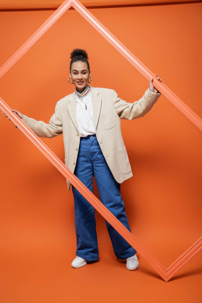 fröhliches afrikanisch-amerikanisches Model in beigem Blazer und blauer Hose mit orangefarbenem Rahmen - Foto, Bild