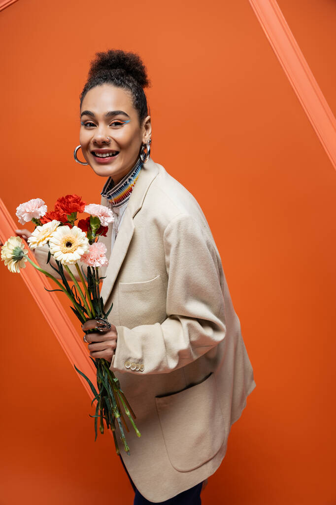 modelo de moda alegre de moda en traje elegante con elegante panecillo agarrando marco naranja y flores - Foto, imagen