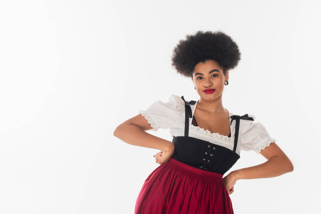 confident african american oktoberfest waitress in traditional attire with hands on hips on white - Photo, Image