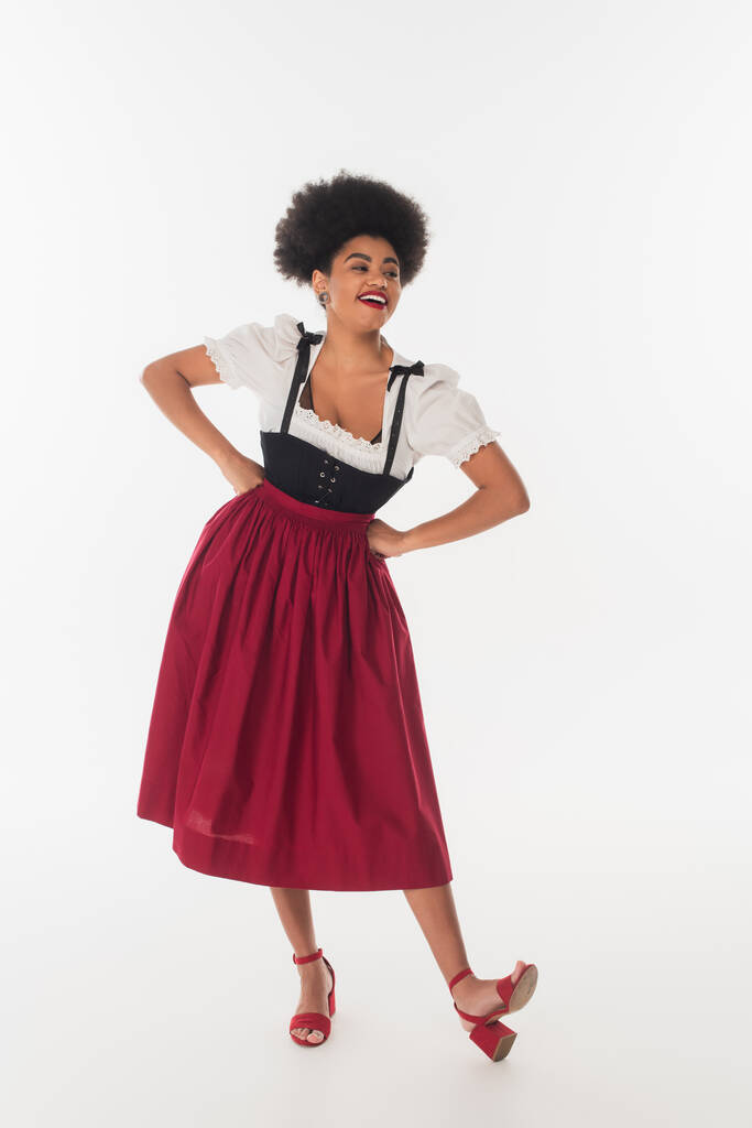 alegre afroamericana oktoberfest camarera en dirndl mirando hacia otro lado con las manos en las caderas en blanco - Foto, Imagen