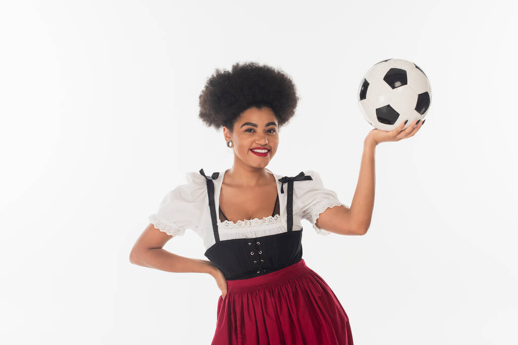glimlachende Afrikaanse Amerikaanse bavariaanse serveerster in dirndl met voetbal op wit, oktoberfest concept - Foto, afbeelding