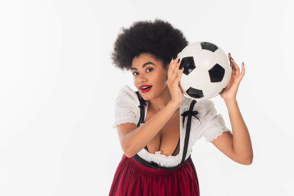 emocionado afroamericano oktoberfest camarera con pelota de fútbol mirando a la cámara en blanco - Foto, Imagen