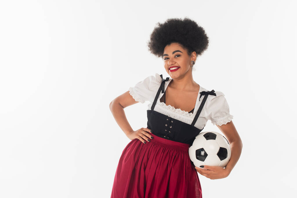 Afrikaans amerikaanse oktoberfest serveerster in bavariaans kostuum met voetbal en hand op de heup op wit - Foto, afbeelding