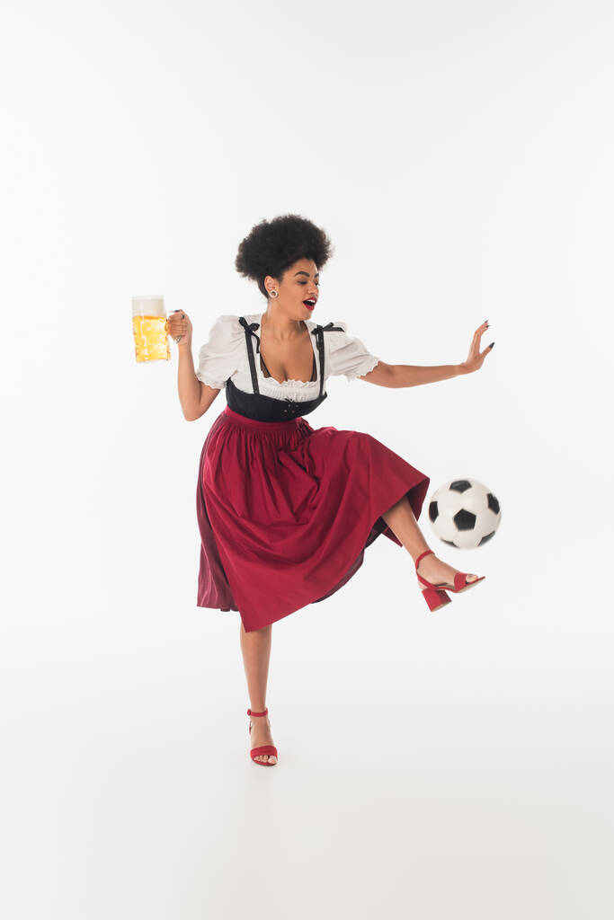 joyeuse serveuse bavaroise afro-américaine avec tasse d'ours mousseux marchant sur le ballon de football sur blanc - Photo, image