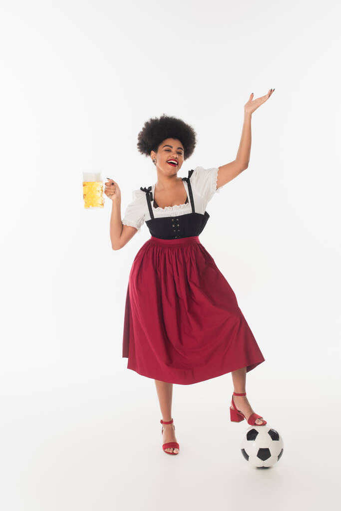 vidám afro-amerikai oktoberfest pincérnő egy bögre kézműves habos sör közelében focilabda fehér - Fotó, kép