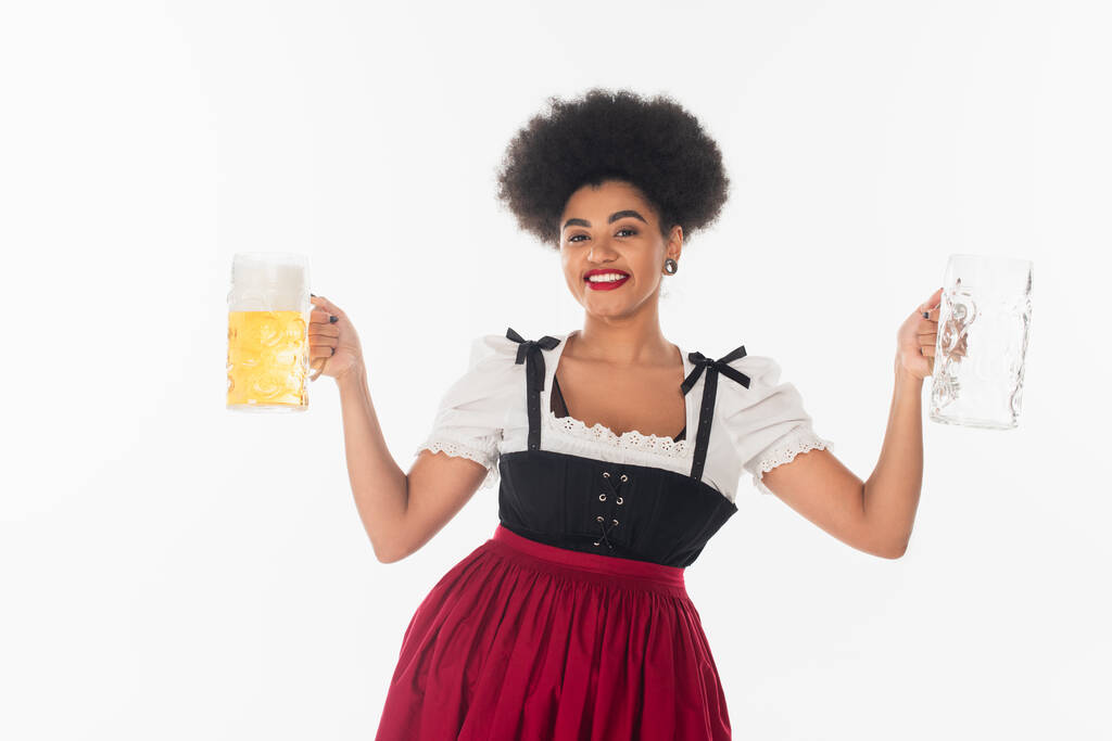 african american bavarian waitress in oktoberfest dirndl with empty and full beer mugs on white - Photo, Image
