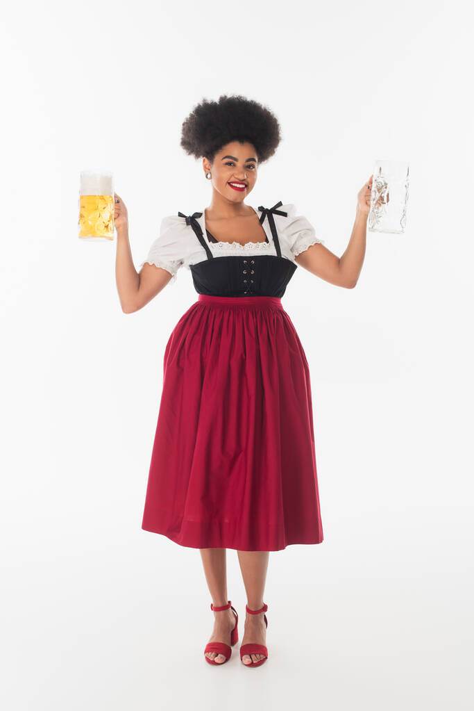 Afrykański amerykański oktoberfest kelnerka w tradycyjnym stroju z pustymi i pełnymi kubkami piwa na białym - Zdjęcie, obraz