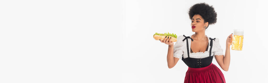 potěšená africká americká bavorská servírka s hrnkem a hot dogem na bílém, oktoberfest, banner - Fotografie, Obrázek
