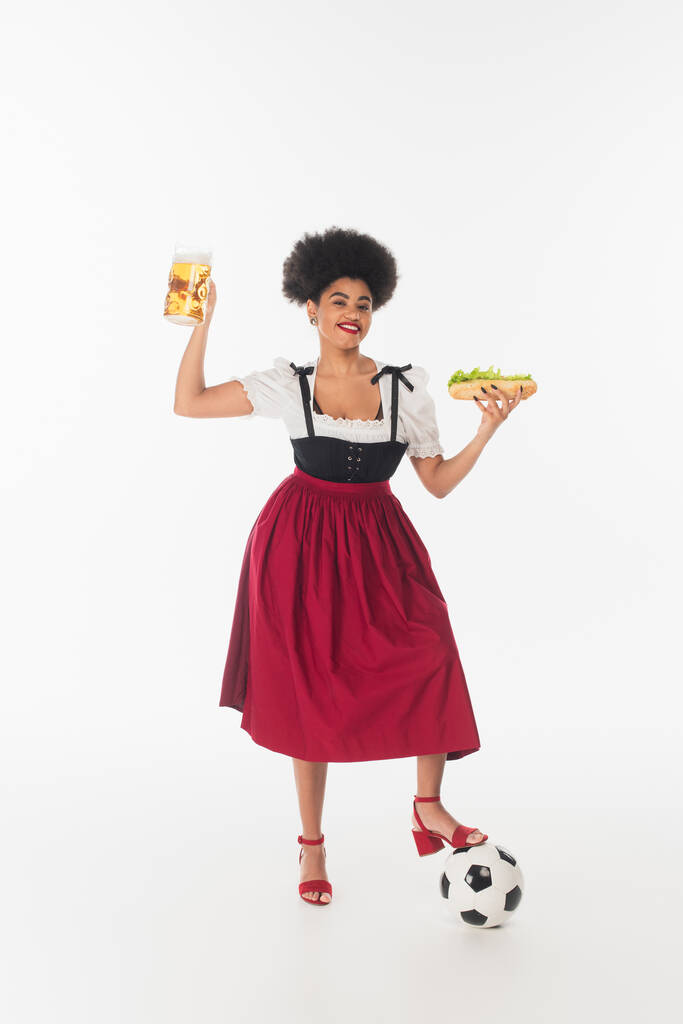 heureuse serveuse bavaroise afro-américaine en dirndl avec tasse de bière, hot dog et ballon de football sur blanc - Photo, image