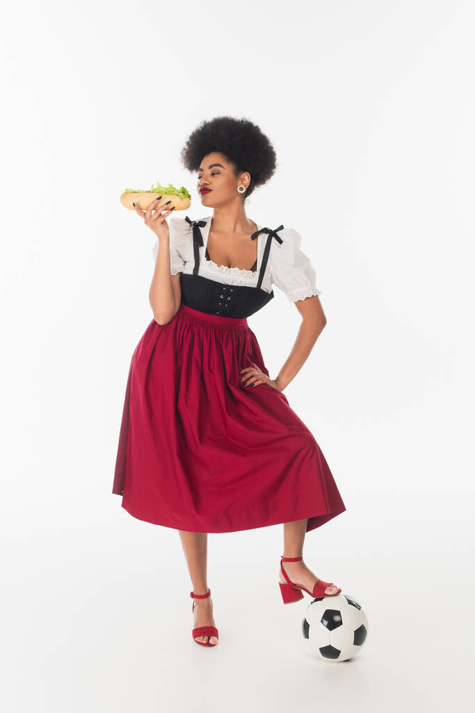 african american bavarian waitress posing with tasty hot dog and soccer ball on white, oktoberfest - Photo, Image