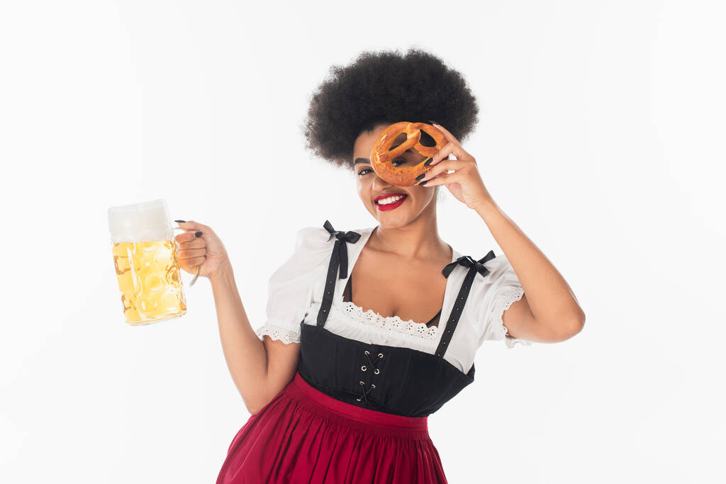 χαρούμενη αφρικανή αμερικανίδα σερβιτόρα Oktoberfest με μια κούπα μπύρα που κρύβει το πρόσωπο της με κουλούρι στα λευκά - Φωτογραφία, εικόνα
