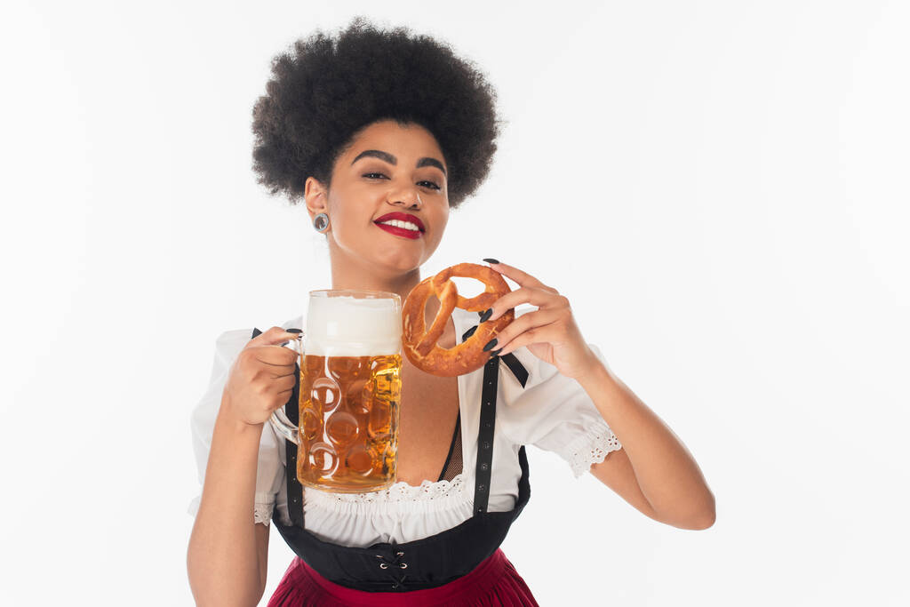 encantada garçonete afro-americana oktoberfest com caneca de cerveja e pretzel olhando para a câmera no branco - Foto, Imagem