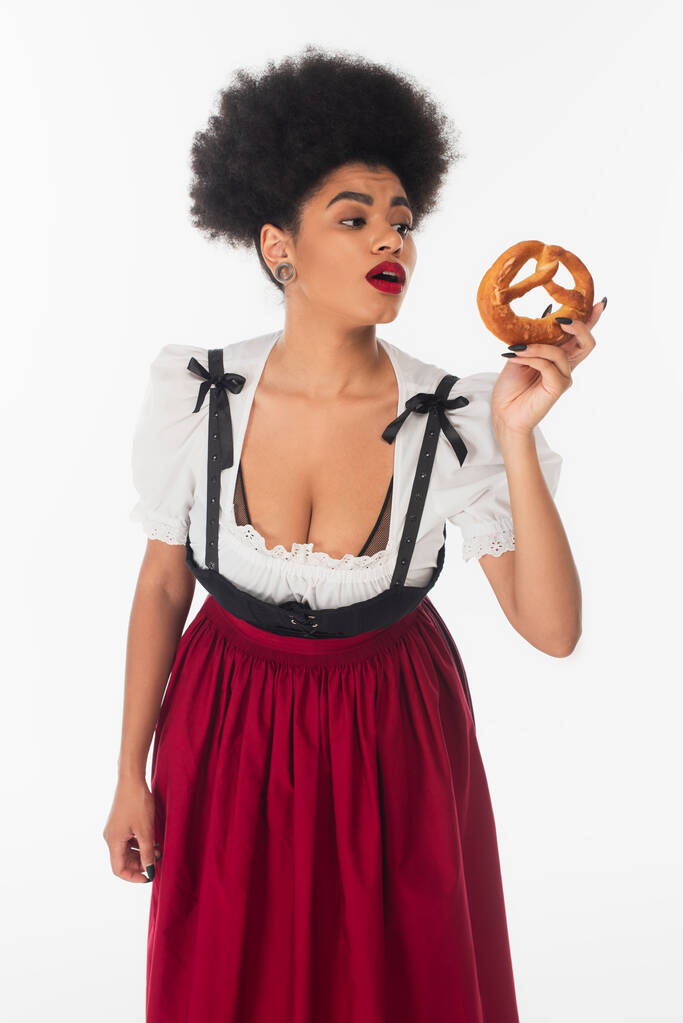 verbaasd afrikaanse amerikaanse bavarian serveerster in oktoberfest kleding op zoek naar smakelijke pretzel op wit - Foto, afbeelding