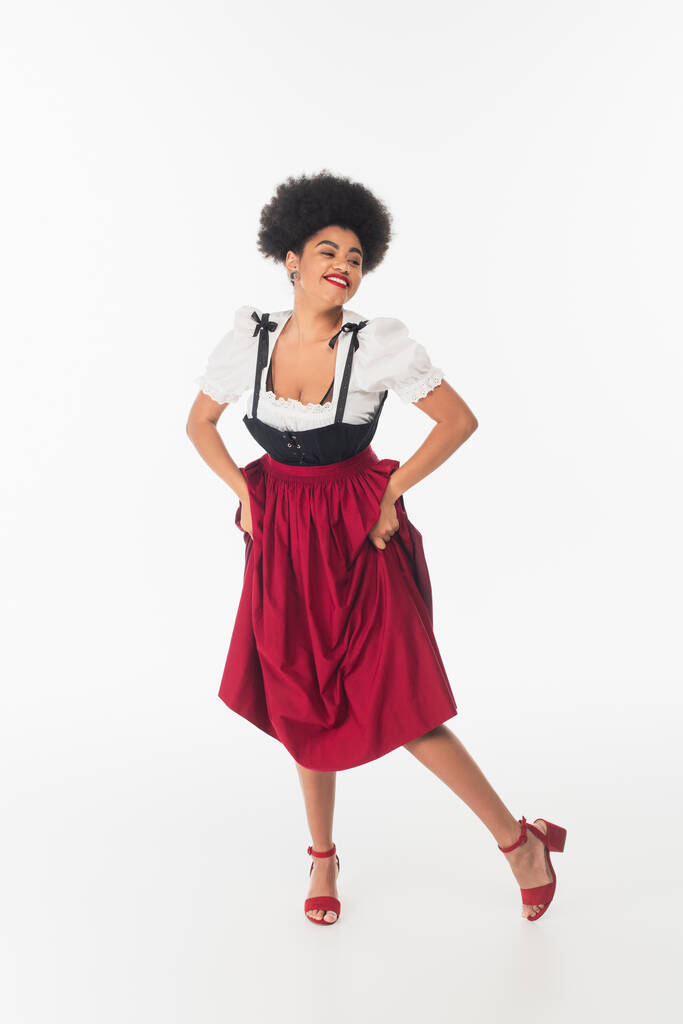 sobregocijado afroamericano oktoberfest camarera en traje bavariano tradicional bailando en blanco - Foto, Imagen