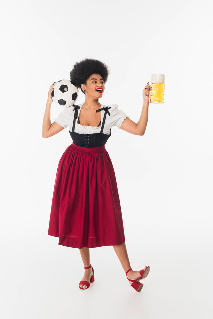 alegre africano americano bavarian camarera en auténtico atuendo con pelota de fútbol y cerveza taza en blanco - Foto, imagen