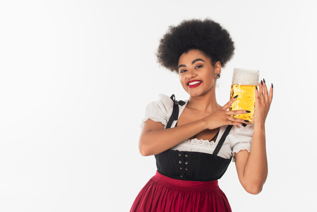 cheerful african american waitress in oktoberfest outfit holding mug of beer with foam on white - Photo, Image