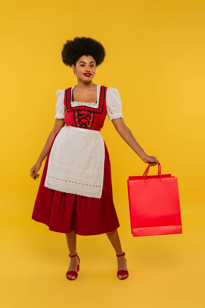 piacere africano americano bavarese cameriera in oktoberfest vestito tenendo shopping bags su giallo - Foto, immagini