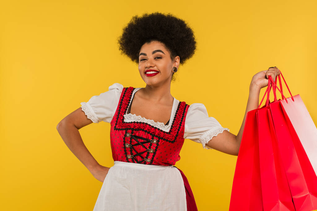 cheerful african american oktoberfest waitress posing with hand on hip and shopping bags on yellow - Photo, Image