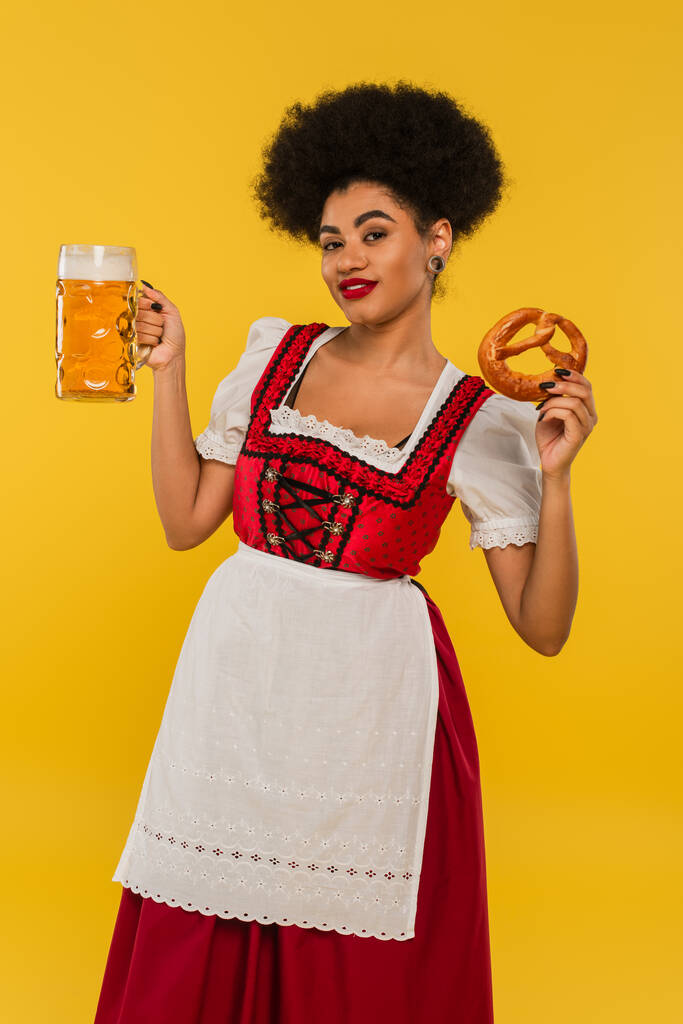 africano americano oktoberfest camarera con taza de cerveza y sabroso pretzel sonriendo a la cámara en amarillo - Foto, imagen