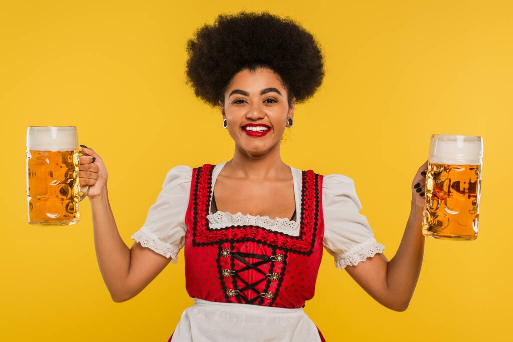 cheerful african american oktoberfest waitress with mugs of craft beer looking at camera on yellow - Photo, Image