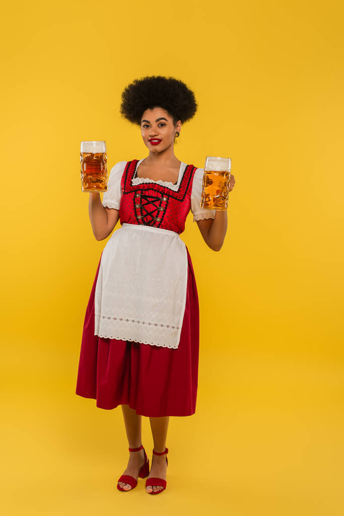 Afrikanisch-amerikanische Kellnerin im Oktoberfest-Dirndl hält Bierkrüge in der Hand und lächelt in die Kamera auf gelb - Foto, Bild