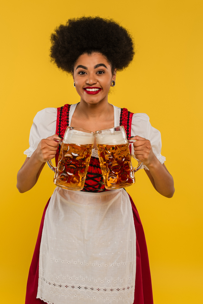 alegre afroamericana camarera en oktoberfest traje tazas de cerveza tintineo y sonriendo en amarillo - Foto, imagen