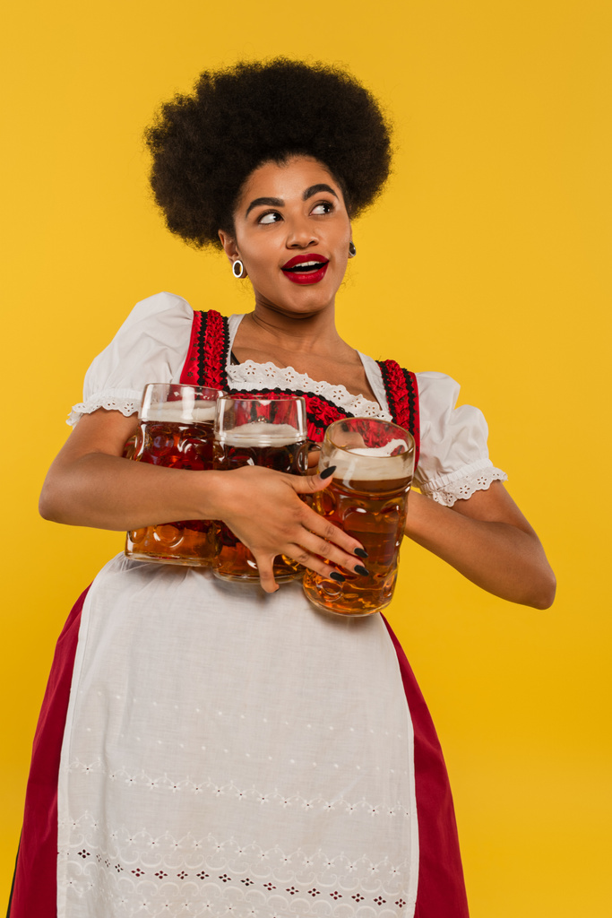 Χαρούμενη Αφροαμερικανή σερβιτόρα στο Ντιρντλ να κρατάει κούπες μπύρας και να κοιτάει αλλού το κίτρινο. - Φωτογραφία, εικόνα