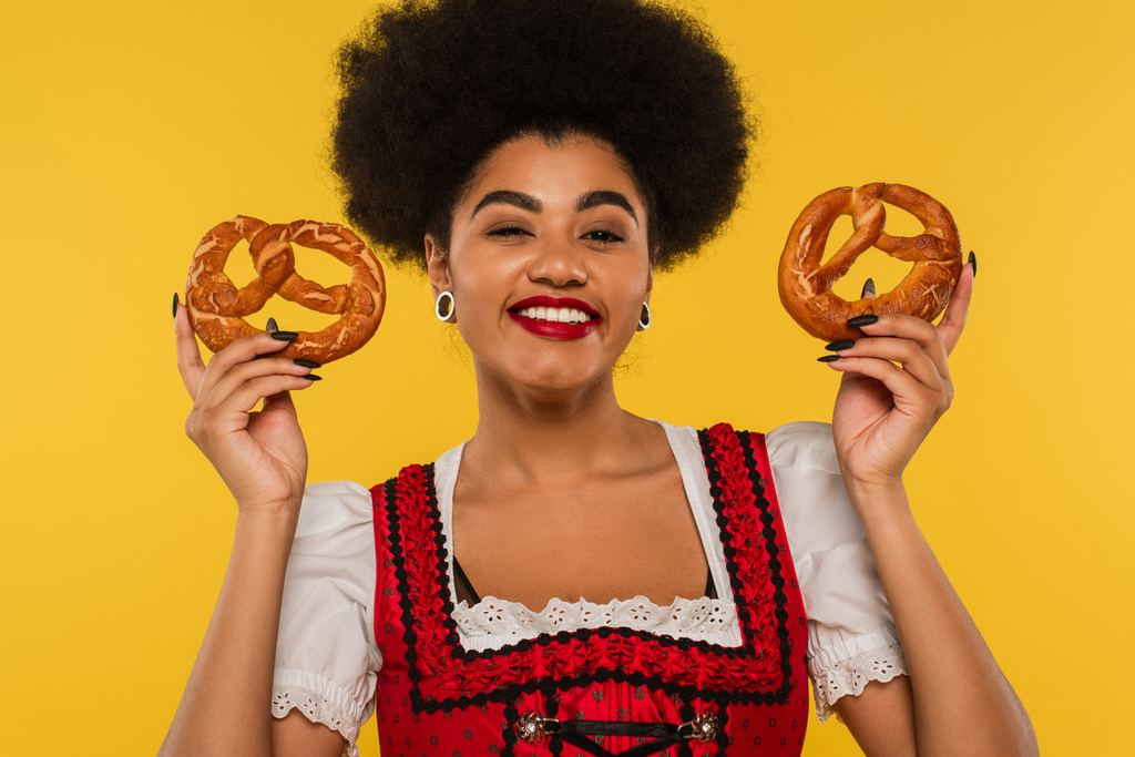 joyeuse serveuse afro-américaine oktoberfest en bavarois dirndl tenant de savoureux bretzels sur jaune - Photo, image